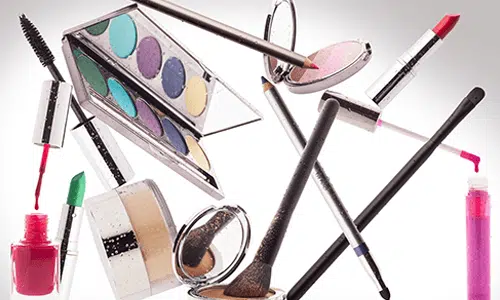 Assorted cosmetics including brushes, palettes, lipstick, and polish floating on a white background